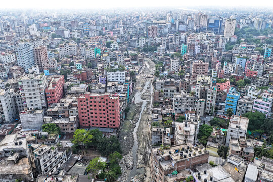 Keraniganj, Bangladesh - 11 April 2026: Aerial view of a sprawling urban tapestry, where a polluted canal snakes through a dense mosaic of grey, white, and red buildings under a hazy sky.