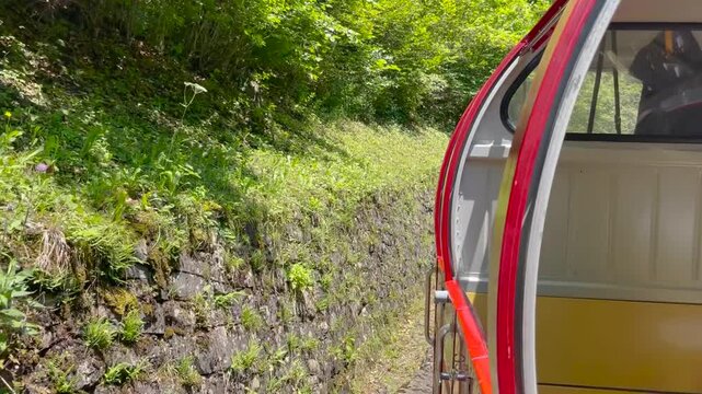 Tracking shot alongside red and yellow steam train passing steep rocky cliff face and lush green forest on Brienz Rothorn Bahn, Switzerland, sunny summer day.