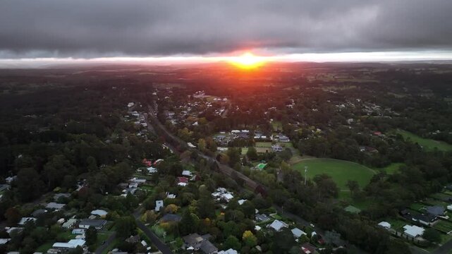 Dramatic low-altitude drone footage of a fiery orange and red sunrise over a green paddock in Bundanoon, Southern Highlands, New South Wales, Australia. Stormy clouds above an intense glowing horizon.