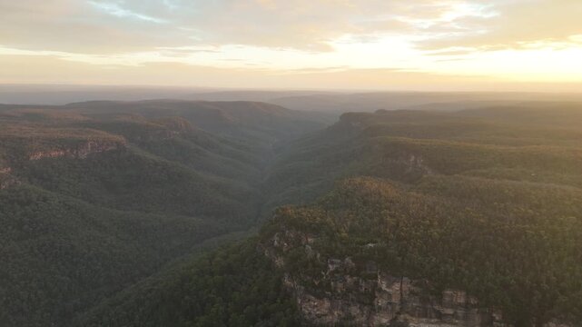 Aerial drone footage rising above a deep forested canyon at sunset in Bundanoon, Southern Highlands, New South Wales, Australia. Golden light glows on the horizon as dark bushland fills the valley bel