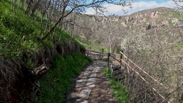 People walk along a narrow stone path surrounded by greenery and trees. The sky is clear, and the sun shines down on the trail. Nature is alive with growth.
