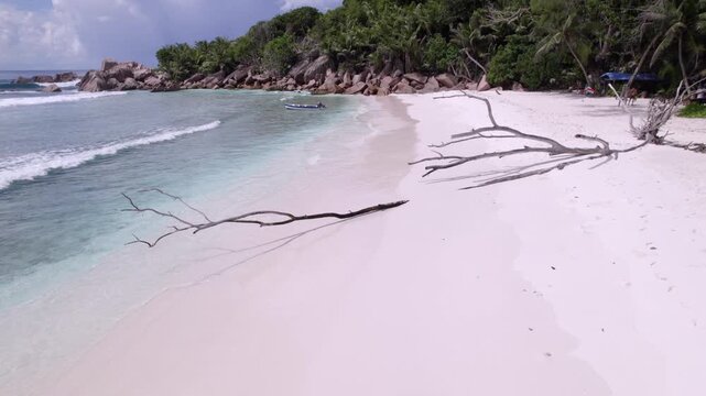 Drone passing near the shoreline near dried tree branch at grand anse beach, granite stones, white sandy beach, docked boat, untouched nature, La Digue, Seychelles 30fps 016.