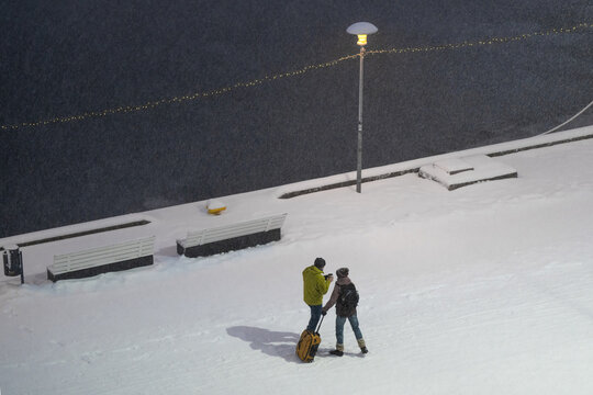 Tallinn, Estonia - 03 January 2025: View of two figures on a snow-white promenade with a yellow suitcase, against dark water and a lone lamp.