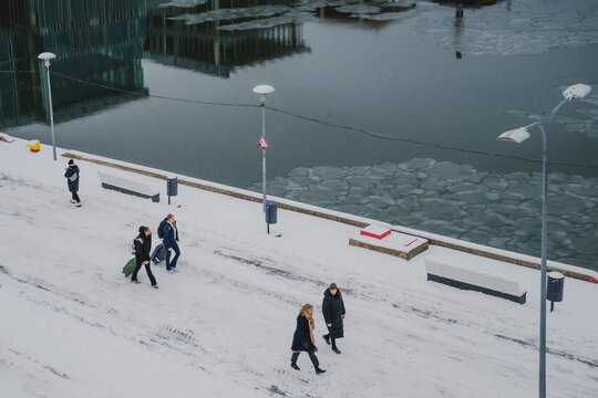 Tallinn, Estonia - 03 January 2025: View of figures walking along the snowy pathway next to the frozen water, reflecting the building's glassy facade under the muted sky.
