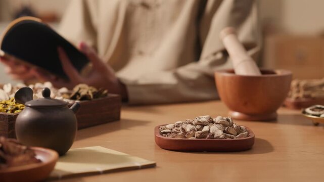 Side pan across dried herbal ingredients in wooden trays while practitioner reads a book. Scene shows study, preparation and use of traditional Chinese medicine materials in apothecary setting.