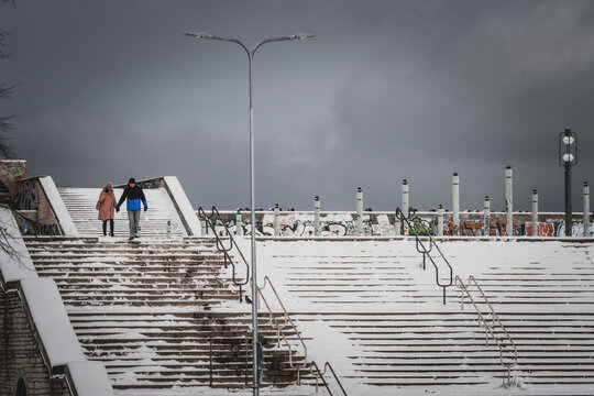 Tallinn, Estonia - 03 January 2025: View of a couple descending a wide, snow-dusted stone staircase under a brooding, overcast sky, contrasting stark whites with dark, textured shadows.