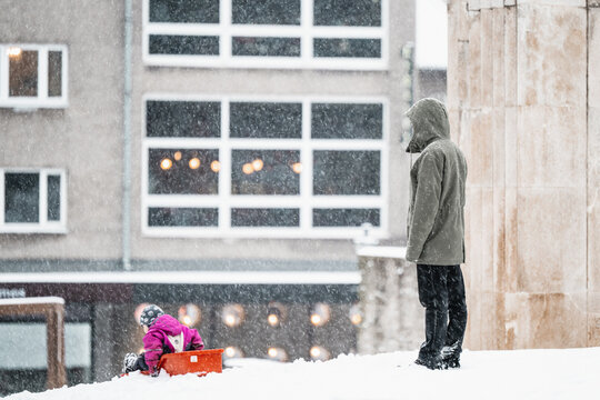 Tallinn, Estonia - 03 January 2025: View of a lone figure watching a child on a vibrant red sled amidst a gentle snowfall and muted urban tones.