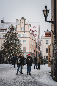Tallinn, Estonia - 03 January 2025: View of snow-dusted streets where people meander past a brightly lit Christmas tree near the historic buildings.