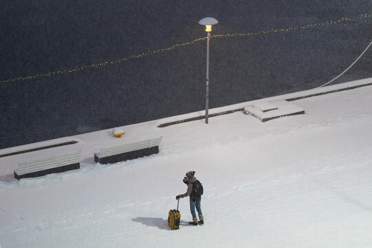 Tallinn, Estonia - 03 January 2025: View of a solitary traveler with a yellow suitcase on a bright, snow-covered pier, illuminated by a streetlamp against the dark water.