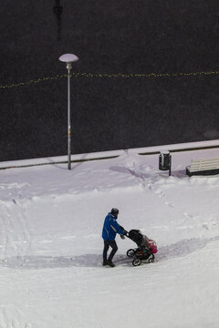 Tallinn, Estonia - 03 January 2025: View of a person pushes a stroller up a snowy slope, the bright white contrasting with the dark background, under a street lamp.