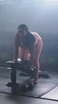 Vertical video: Gripping dumbbell, trainee doing one-arm row at gym bench for strength, in pink