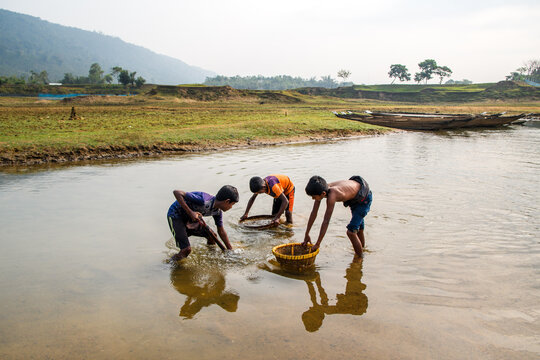 Sylhet, Bangladesh - 11 December 2019: View of children wading in shallow water, their reflections shimmering as they sift through sand with baskets, framed by distant hills.