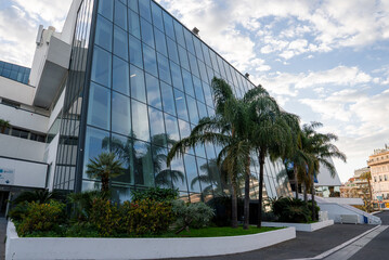 Obraz premium Palais des Festivals et des Congres in Cannes, France stands with a sharply slanted facade, palm trees, curving promenade, white terraced ramps, and soft daylight skies.