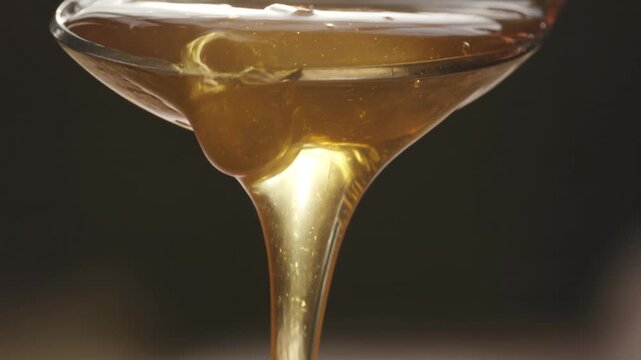 Golden honey flows smoothly from a spoon into a glass dish, showcasing its rich texture and viscosity against a dark background in a close-up shot