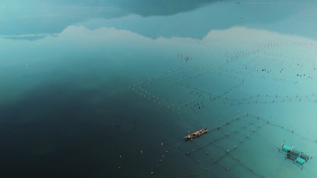 Fishing boat gliding across cau hai lagoon amid geometric aquaculture nets and traps, sky reflections on calm blue water evoking traditional vietnamese coastal livelihood and serenity