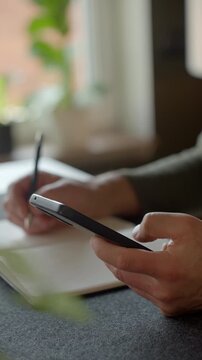 Man checking smartphone while writing notes in notebook at home desk near laptop. Concept of planning, productivity and using technology in everyday life