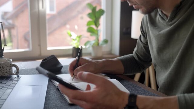 Close up of man writing notes in notebook while using smartphone at desk in home office. Concept of planning, mobile communication, productivity and remote work