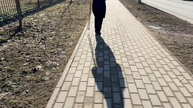 Woman Nordic walking shaded park path, brisk stride with poles and long shadow cast on paving, lea strewn verge, cool crisp light