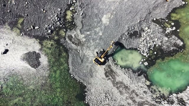 Heavy machinery works in a granite quarry located next to water pools. The scene shows rock removal and transportation activities under bright sunlight.
