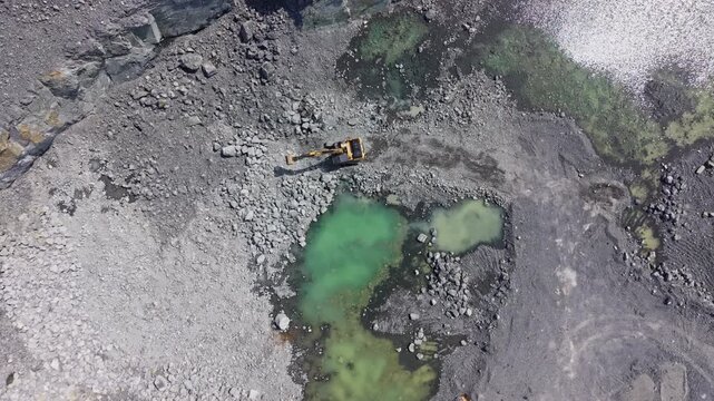 Excavation is ongoing in a granite quarry. Heavy machinery is moving stones and gravel. There is a body of green water in the quarry area. The scene shows raw terrain and rocky surfaces.