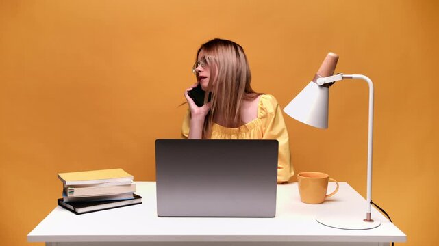 Young businesswoman talking on a smartphone while working on a laptop at her office desk against a yellow background