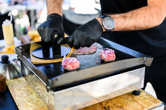 Chef preparing smash burgers on a flat top grill, adding toppings to freshly cooked beef patties with melted cheese, capturing street food cooking process and professional burger preparation in a