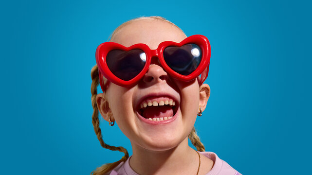 Child girl wearing heart sunglasses laughing on blue background with bright studio lighting and tight crop. Concept of happiness, kids lifestyle, social media and advertising visuals.