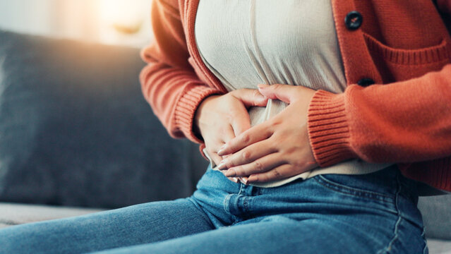 Cramps, menstruation and hands of woman on sofa in home for period pain, healthcare and stomach virus. Endometriosis, constipation and bloating with person in living room of house for pms or diarrhea