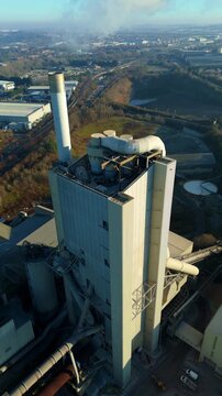 Vertical drone shot looking up toward the smoking chimney stack of a British cement factory.