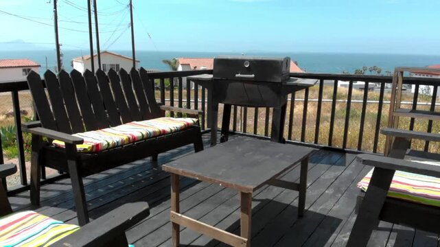 Wooden deck with outdoor seating, table and grill, cushions on chairs and ocean visible in background, relaxed coastal setting showing leisure, comfort and vacation lifestyle