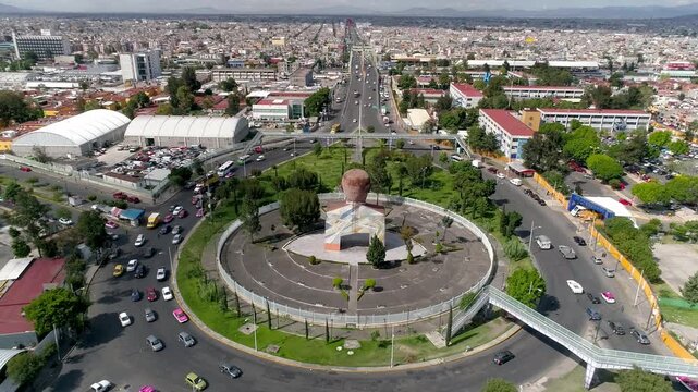 Aerial drone shot of roundabout with monument at center, vehicles moving along circular roads and highway stretching across city, showing traffic flow and urban layout