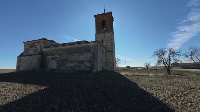 Slow motion pullback at 24fps of a ruined church in backlight, showing tower and nave silhouette with a cement blocked entrance and its large shadow cast over plowed soil.