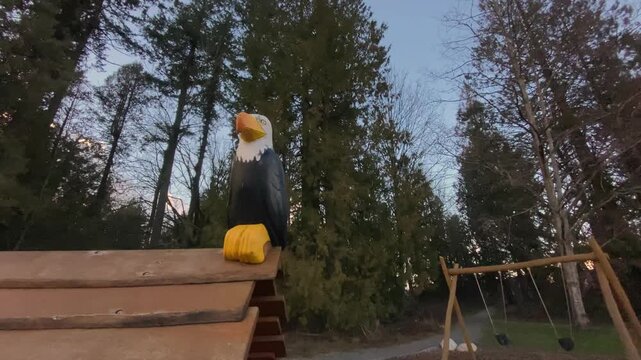 Bald eagle wooden carving perched on play structure roof viewed from low vantage point slowly moving backward revealing empty swings hanging over wood chips during suburban sunset