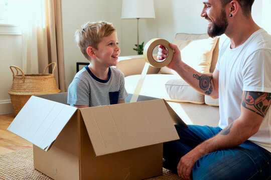 Caucasian father and young son packing cardboard box with tape, smiling during home move and family organization. Ideal for relocation services, parenting, storage, moving day marketing