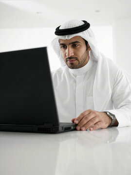 Low angle Saudi businessman with shemagh and agal using laptop in minimal white setting