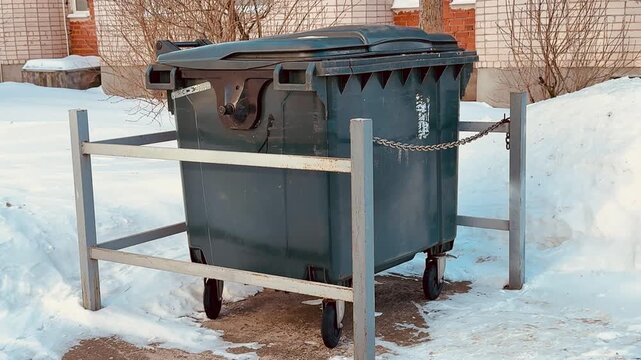 Plastic trash can behind fence, snow. Blue gray plastic dumpster chained to metal rail beside brick wall, snowbanks piled