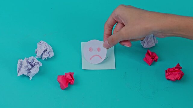 Slow motion flatlay of Hand covering smile with  frowning emoji on turquoise background