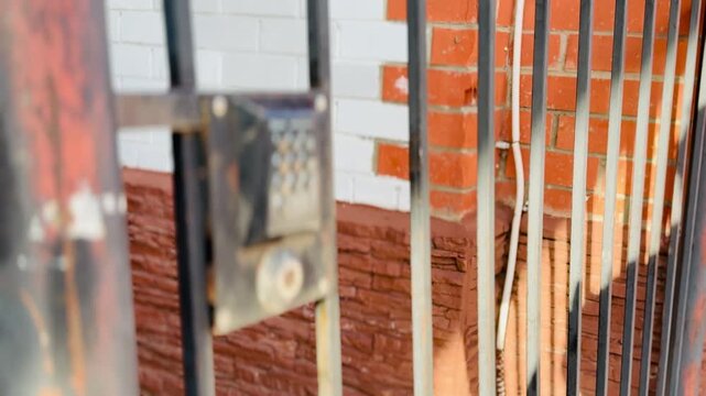 Close-up black intercom on fence, unfocused daytime street scene with rusty metal bars and red brick wall, textured paint chips