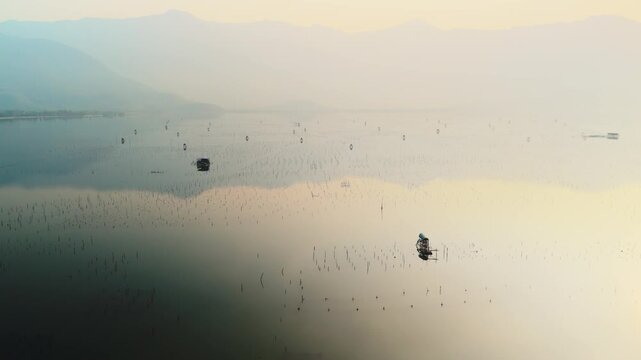 Fisherman working on an oyster farm at sunrise, his small boat gently moving across the calm, reflective waters of lap an lagoon, framed by distant misty mountains under a soft sky