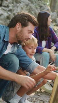 Father and son making together campfire enjoy spend camping evening time outdoors on nature. Smiling mother and daughter are preparing food for a picnic on background. Children with parents are