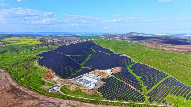A vast area shows rows of solar panels set on green land. Workers are present near a site that appears to be under construction. The sky is clear, and hills are visible in the background.