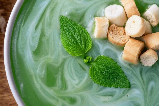 Close up of a bowl of mint or nettle leaves green soup sits on a wooden table. The soup is swirled with cream and topped with croutons. top view. Healthy food