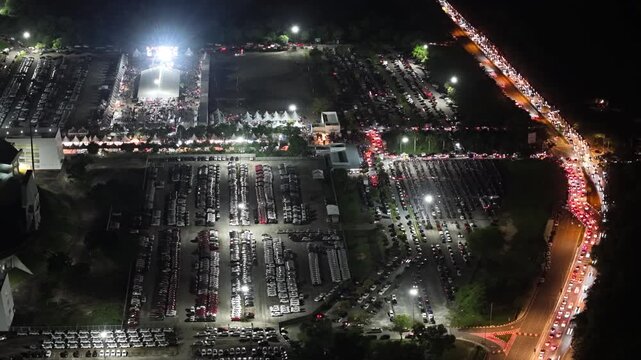 Aerial night footage of a packed parking lot and long vehicle light trail on adjacent road at Batu Kawan Stadium