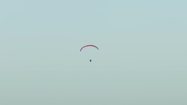 Colorful paraglider wing carrying paramotor pilot cruising across pale blue sky minimal background. Powered paragliding flight moving smoothly through open air extreme outdoor sport energy. Solo