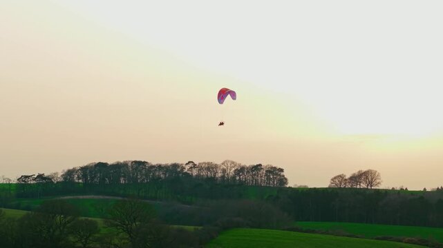 Colorful powered paraglider flying across golden dusk sky above dark woodland horizon countryside panorama. Paramotoring pilot cruising through calm evening air extreme outdoor activity adventure