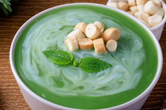 Close up of a bowl of mint or nettle leaves green soup sits on a wooden table. The soup is swirled with cream and topped with croutons. top view. Healthy food