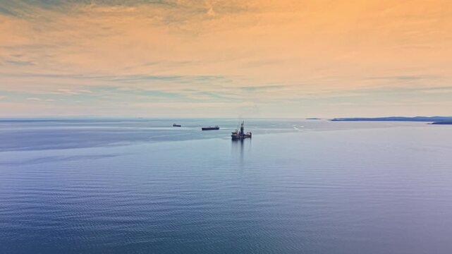 An oil tanker travels through the open sea near a drilling rig at sunset. Other ships are visible on the horizon. The sky shows warm colors as the day ends.
