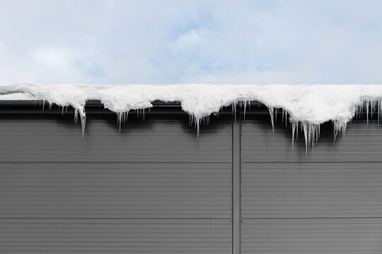 Thick layer of white snow and icicles hang from the edge of a grey metal roof against the sky.