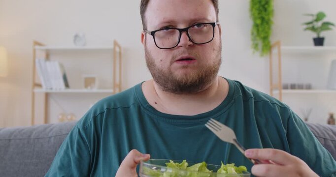 At home, fat overweight man in glasses eats a salad with a fork from a bowl by the window, suggesting a healthy lunch, nutrition and diet in a bright kitchen. Simple healthy meal. Clean life.