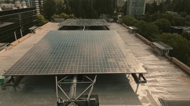Large solar panel array reflecting bright afternoon sunlight powering smart hydrogen facility mounted on concrete parking garage roof surrounded by trees captured via retreating low angle drone
